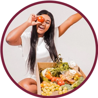 Person smiling, holding a tomato, seated beside a box of fresh vegetables and fruit
